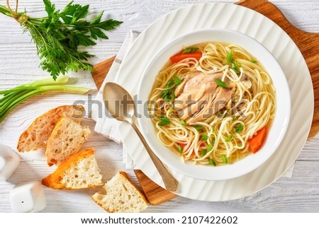chicken noodle soup with carrots and scallion in a white bowl on a white wooden table with white bread and spoon, horizontal view from above, flat lay, close-up