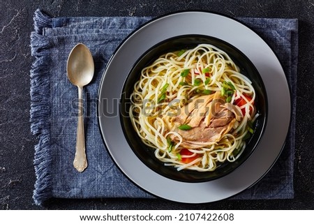 chicken noodle soup with carrots and scallion in a black bowl on a grey concrete table with spoon, horizontal view from above, flat lay, close-up