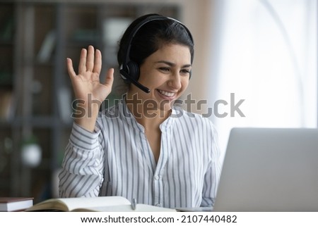 Happy attractive millennial Indian ethnicity woman in headphones with microphone waving hand, starting web camera video call meeting with colleagues or teacher. distant communication concept. Royalty-Free Stock Photo #2107440482