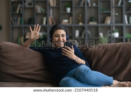Smiling attractive millennial Indian woman in headphones looking at cellphone screen, involved in pleasant web camera call conversation chatting with friends, talking discussing life news distantly. Royalty-Free Stock Photo #2107440518
