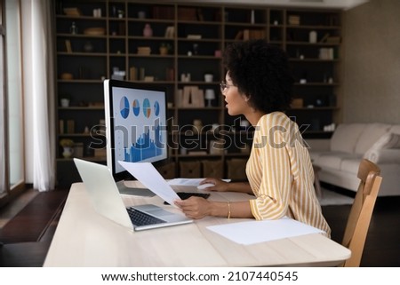 Happy young African American businesswoman in eyeglasses looking at computer monitor, analyzing online sales or marketing research graphs using modern technology gadgets, working with statistics. Royalty-Free Stock Photo #2107440545