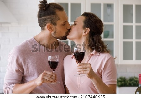 Affectionate loving young man woman family couple kissing, clinking glasses with red wine, enjoying romantic home date or celebrating wedding anniversary or special occasion, spending time together.