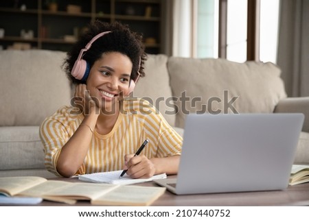 Happy young pretty African American woman in headphones looking at laptop screen, writing notes watching educational lecture, improving knowledge distantly, preparing for exams or studying remotely. Royalty-Free Stock Photo #2107440752