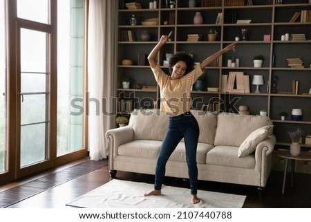 Happy carefree young African American woman dancing on floor carpet, listening energetic disco in living room, laughing enjoying hobby leisure time, celebrating freedom or moving into own home. Royalty-Free Stock Photo #2107440788