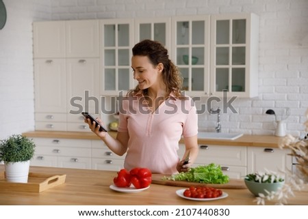 Happy beautiful young Caucasian woman using cellphone, web surfing interesting tasty recipe or sharing meal preparing process, enjoying cooking healthy vegetarian began food alone in modern kitchen.
