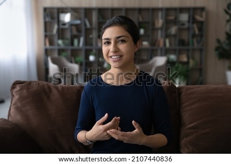 Attractive happy young Indian woman talking holding web camera call, sitting on comfortable sofa at home. Smiling female blogger recording educational video, distant communication concept. Royalty-Free Stock Photo #2107440836