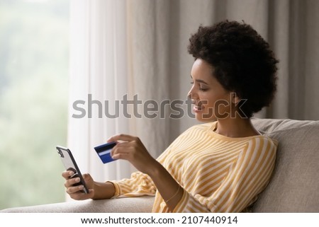 Happy young African American woman holding cellphone and bank credit card in hands, transferring money online, shopping goods in internet store, purchasing services, satisfied with secure payments. Royalty-Free Stock Photo #2107440914