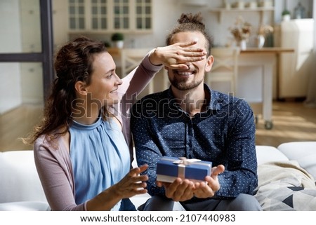Caring cheerful young woman covering eyes of excited husband, giving prepared gift box making surprise, happy birthday congratulation, wedding anniversary or special event celebration concept.