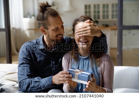 Smiling millennial hipster man covering eyes of joyful curious young wife, giving wrapped box with gift, preparing surprise congratulating with happy birthday, marriage anniversary, special occasion.