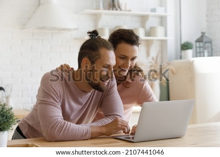 Smiling bonding young family couple looking at laptop screen, watching entertaining photo video content online, web surfing information, choosing goods shopping in internet store, booking tickets.