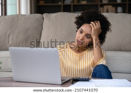 Unhappy thoughtful young African American woman looking at computer screen, feeling stressed managing household budget, suffering from lack of money paying for utility bills in e-banking application.
