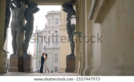 Beautiful newlyweds hug at old building. Action. Small silhouette of newlyweds in distance under arch of tall statues. Beautiful shot with tall statues under arch of building and newlyweds Royalty-Free Stock Photo #2107447973