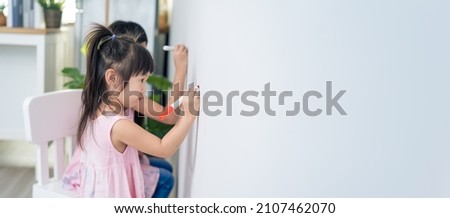 Asian young sibling kid girl enjoy paint on white wall in living room. Little adorable children having fun drawing and coloring art picture with hapiness enjoy creativity activity on holiday at home.