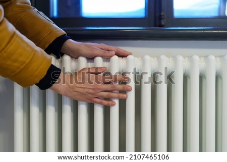 Cropped photo of woman warming hands near radiator at home after walking in cold winter weather, female touching barely warm battery during heating season, person near window checking heating system