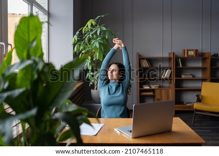 Relaxed office worker woman stretching hands and body taking break from work on laptop smiling look in window. Joyful freelancer copywriter girl happy with task done at workplace in coworking space Royalty-Free Stock Photo #2107465118
