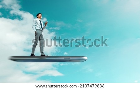 User Experiences for Mobile Concepts. Smiling Young Businessman Standing on Huge Flying Smartphone while Using Mobile Phone in the Sky. Manipulation Photo. Low Angle View Royalty-Free Stock Photo #2107479836