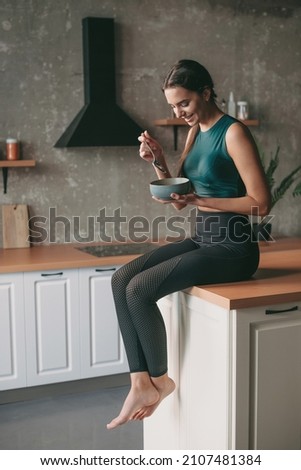 Caucasian woman in sportswear resting at the kitchen after exercise and eating a bowl of porridge. Vegetarian healthy food. Royalty-Free Stock Photo #2107481384