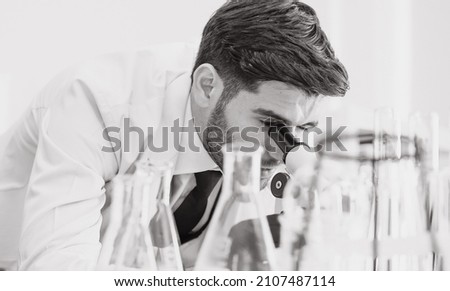 Bearded man scientist looks through microscope in a medical science research laboratory, portrait. Doctor analyzing innovative virus protective vaccines in health care lab. Black and white photograph