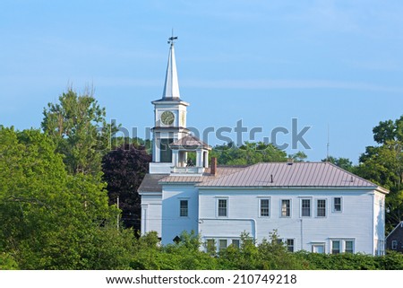 A church with clock and weathervane upon the spire in the background with a building with a bell tower in the foreground in Frankfort, Maine.