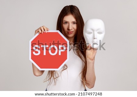 Portrait of serious dark haired woman holding white mask with unknown face and red traffic sign, looking at camera, wearing white T-shirt. Indoor studio shot isolated on gray background.