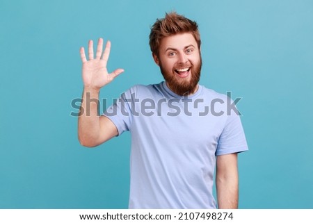 Portrait of positive bearded man greeting you rising hand and waving, saying hi, glad to see you, looking at camera with toothy smile. Indoor studio shot isolated on blue background. Royalty-Free Stock Photo #2107498274