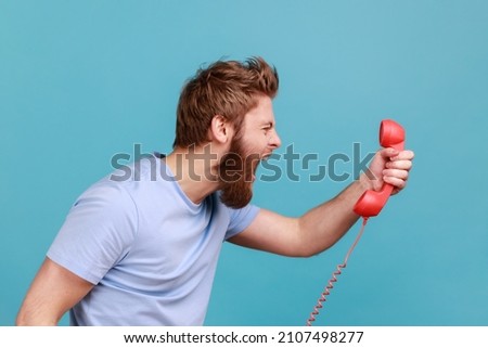 Side view portrait of handsome angry bearded man screaming and yelling talking retro landline phone, complaining on connection quality. Indoor studio shot isolated on blue background. Royalty-Free Stock Photo #2107498277