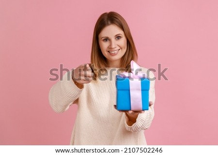 Satisfied young adult blond woman standing with blue gift box in hands and pointing finger to camera, choosing you as winner, wearing white sweater. Indoor studio shot isolated on pink background.