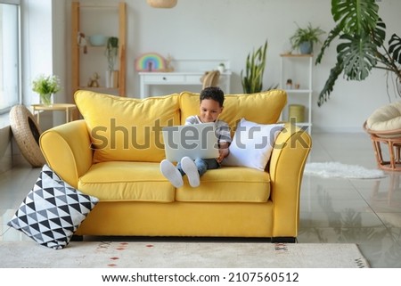 Little African-American boy with laptop watching cartoons on sofa at home