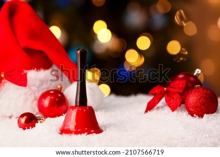 Christmas bell, decorations and Santa hat on snow against blurred lights
