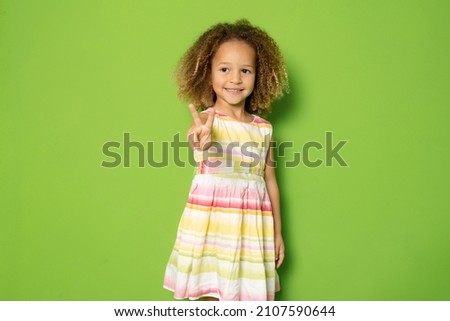 Full length photo of excited charming cute little girl showing peace symbol standing isolated over green background.