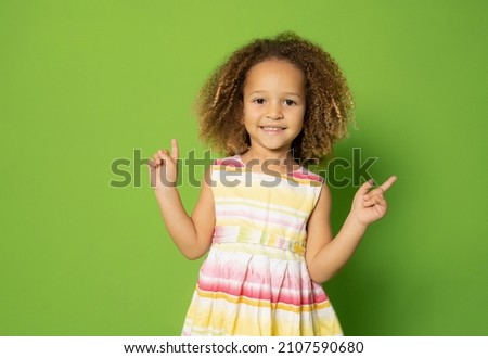 Little kid girl 3-4 years old in dress point fingers up on workspace commercial promo area mockup copy space isolated on green background Royalty-Free Stock Photo #2107590680