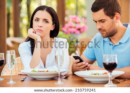 This is the worst date ever. Depressed young woman holding hand on chin and looking at camera while her boyfriend talking on the mobile phone at outdoors restaurant Royalty-Free Stock Photo #210759241
