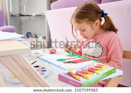 Little girl sits by desk and paints a picture with acrylic paints.