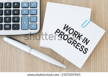 a white card with the text WORK IN PROGRESS on a wooden table next to a pen and calculator
