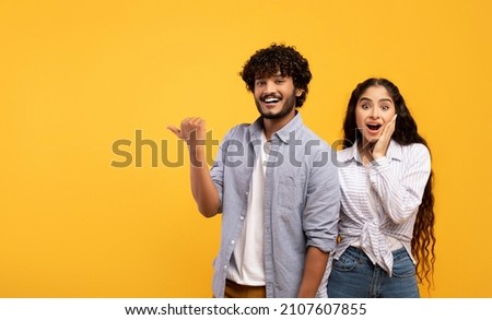 Omg, check this. Emotional indian guy pointing finger aside at free space over yellow background, surprised lady looking at camera, panorama. Couple demonstrating place for your text Royalty-Free Stock Photo #2107607855