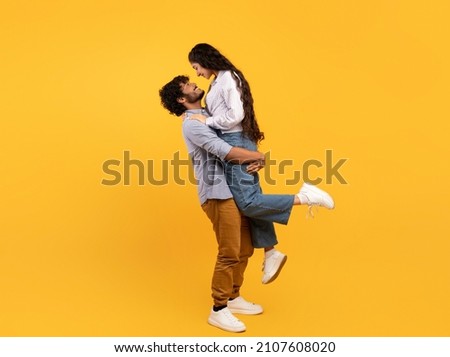 Loving indian guy lifting his girlfriend and holding her on yellow studio background, profile side view, free space banner. Romantic couple expressing their love to each other