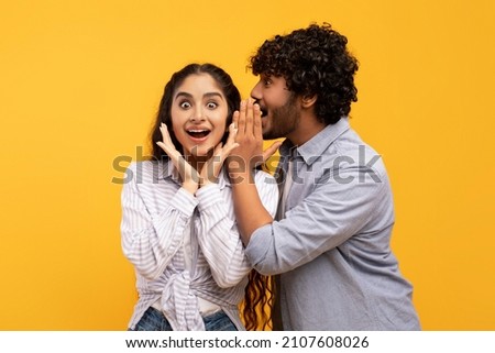 Shock, gossip, share advice. Young indian man whispering to lady on ear, happy woman with open mouth excited and surprised, posing on yellow studio background Royalty-Free Stock Photo #2107608026