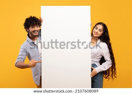 Happy indian couple in love standing by white empty board for advertisement or text over yellow studio background. Excited man and woman standing next to blank placard for ad Royalty-Free Stock Photo #2107608089