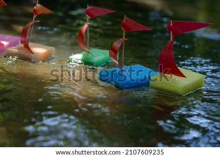Children's ships in the summer river. Homemade boats from kitchen sponges with masts. Mock sports regatta. Shooting at water level. Selective focus. Blurred motion.