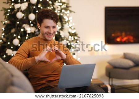 Love And Care. Portrait of smiling man in wireless headphones making video conference with beloved girlfriend, showing heart sign gesture with hands to webcam. Guy sitting on couch near fireplace