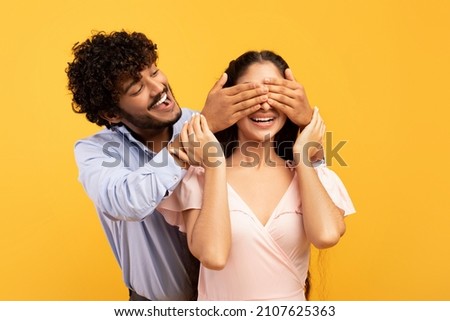 Romantic surprise concept. Portrait of happy indian guy covering his pretty girlfriend eyes from back, standing behind her, couple posing on yellow studio background, banner Royalty-Free Stock Photo #2107625363