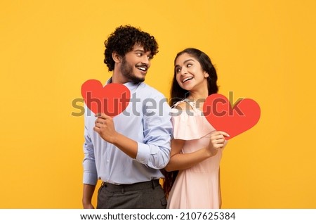 Valentine's holiday. Portrait of indian lovers holding red paper hearts and looking at each other, standing on yellow studio background. Romantic relationship concept Royalty-Free Stock Photo #2107625384