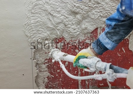 Plasterer operating sprayer equipment machine for spraying thin-layer putty plaster finishing on on a concrete wall covered with a red primer Royalty-Free Stock Photo #2107637363