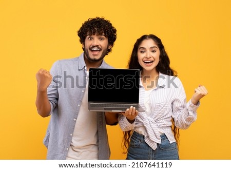 Yes. Portrait of happy indian couple holding laptop with blank screen and shaking clenched fists, showing computer with empty free space for mockup, yellow studio background