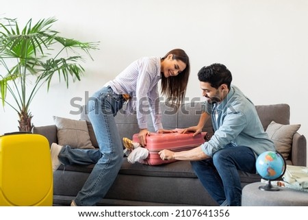 Preparation for vacation. Happy spouses packing and trying to close suitcase on sofa, preparing for travel at home. Vacation, family travel together and positive emotions Royalty-Free Stock Photo #2107641356