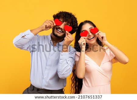 Lovers blinded by their big love. Young indian couple in love holding red heart-shaped cards over eyes and smiling, standing over yellow studio background. St. Valentines Day celebration concept Royalty-Free Stock Photo #2107651337