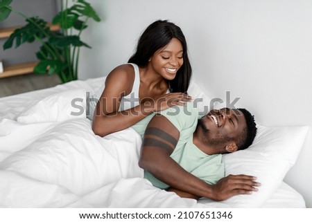 Smiling young pretty african american woman wakes up her husband after night sleep on bed in bedroom. Wake up and good morning, sleep together, love and relationship at home. Couple enjoy free time Royalty-Free Stock Photo #2107651346