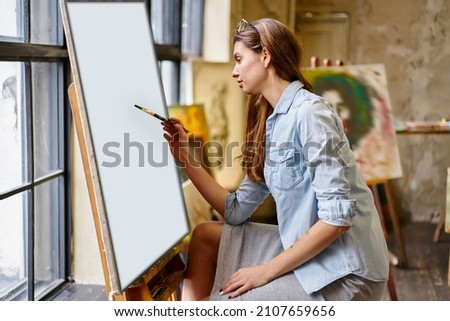 Casual dressed female artist with paintbrush drawing abstract paintings on canvas have workshop at easel, talented Caucasian mastery creating picture during weekend inspiration in art studio
