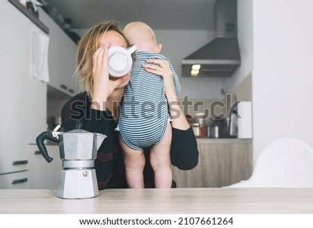 Modern young tired mom and little child after sleepless night. Exhausted woman with baby is sitting with coffee in kitchen. Life of working mother with baby. Postpartum depression on maternity leave. Royalty-Free Stock Photo #2107661264