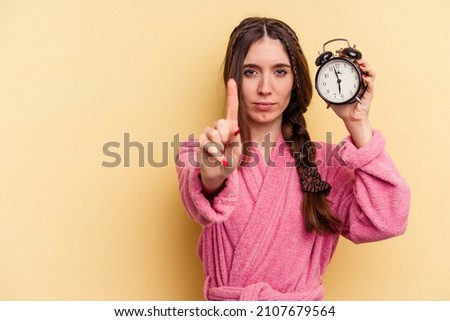 Young caucasian woman wearing a bathrobe holding a alarm clock isolated on yellow background showing number one with finger. Royalty-Free Stock Photo #2107679564